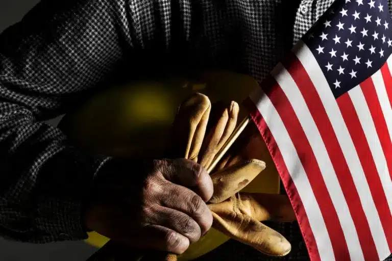 A person holds a small American flag, brown leather gloves, and a yellow construction helmet while wearing a checkered shirt.